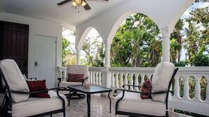 A covered patio with two white cushioned chairs, a small table, and a ceiling fan. The space is enclosed by white columns and features a view of lush greenery. - R Casa Unit 3 At Steps Beach (Rincon)