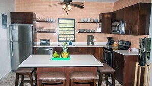 A compact kitchen with a refrigerator, microwave, and stove. There are two stools at the island, and a ceiling fan above. - R Casa Unit 3 At Steps Beach (Rincon)