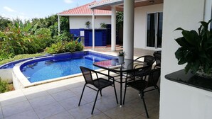 A covered outdoor area with a swimming pool, a table with chairs, and potted plants.