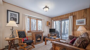 A cozy living room with a fireplace, wooden furniture, and large windows offering a view of trees. - RMR: Sagebrush 1311 Condo in The Aspens/Wilson,WY (Wilson)