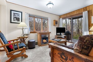 A cozy living room with a fireplace, wooden furniture, a television, and large windows offering a view of trees.