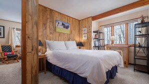 A bedroom with a wooden wall panel, a bed with white bedding, a wooden chair, a small table with a lamp, and a bookshelf. - RMR: Sagebrush 1311 Condo in The Aspens/Wilson,WY (Wilson)