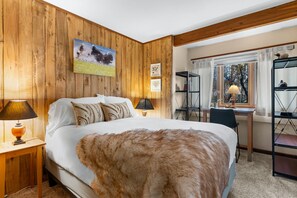 A bedroom with a wooden wall, a bed with a white comforter, a fur throw, a desk with a lamp, and a window with curtains.
