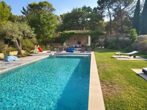 Une piscine rectangulaire à l’eau d’un bleu limpide, entourée d’une pelouse où sont disposés des chaises longues et un petit bâtiment doté d’un toit en tuiles.