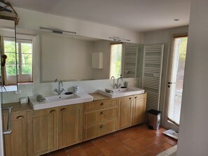 Une salle de bain moderne avec deux vasques, des meubles en bois, un grand miroir et une fenêtre avec des stores.