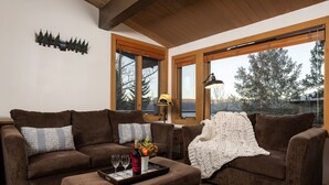 A cozy living room with two brown sofas, a coffee table with a bottle and glasses, and a large window with a view of trees. - Aspenwood L10 - Walk to Slopes | Cozy Mountain + Hot Tub (Snowmass Village)