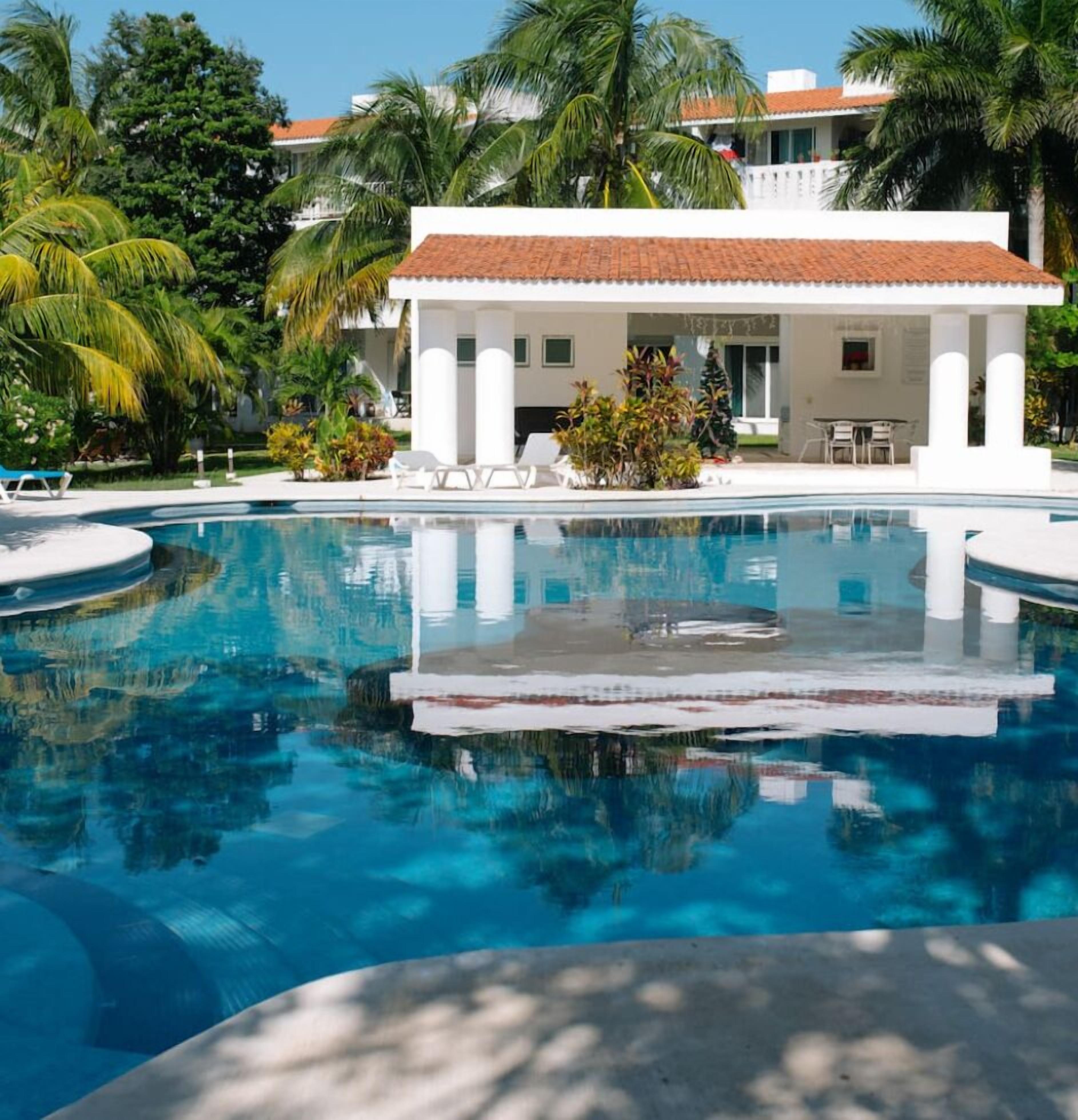A clear blue pool with a white building in the background, surrounded by palm trees.
