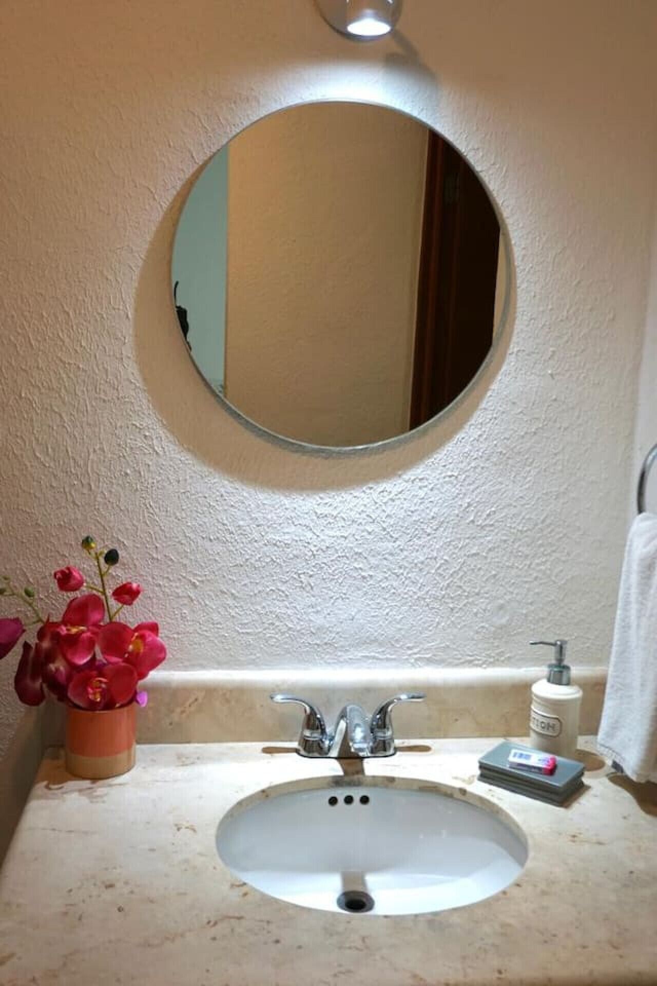 A bathroom with a round mirror, a sink, a flower vase, and toiletries.