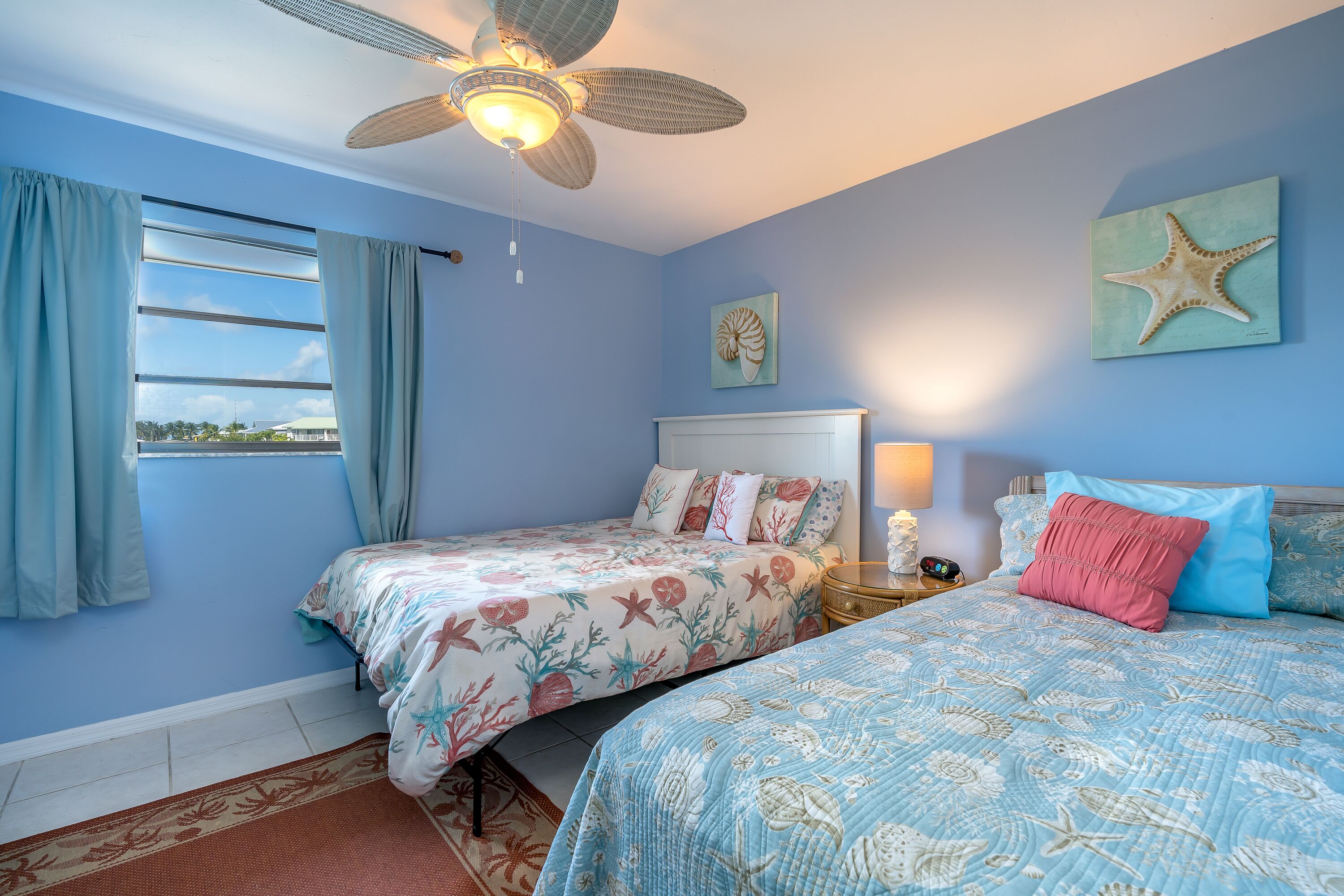 A bedroom with two beds, a ceiling fan, a window with curtains, and a starfish artwork on the wall.