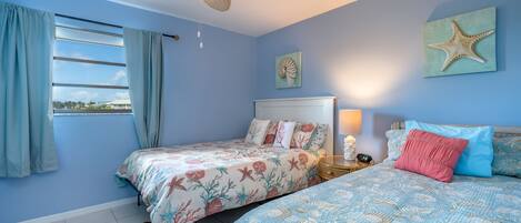 A bedroom with two beds, a ceiling fan, a window with curtains, and a starfish artwork on the wall.