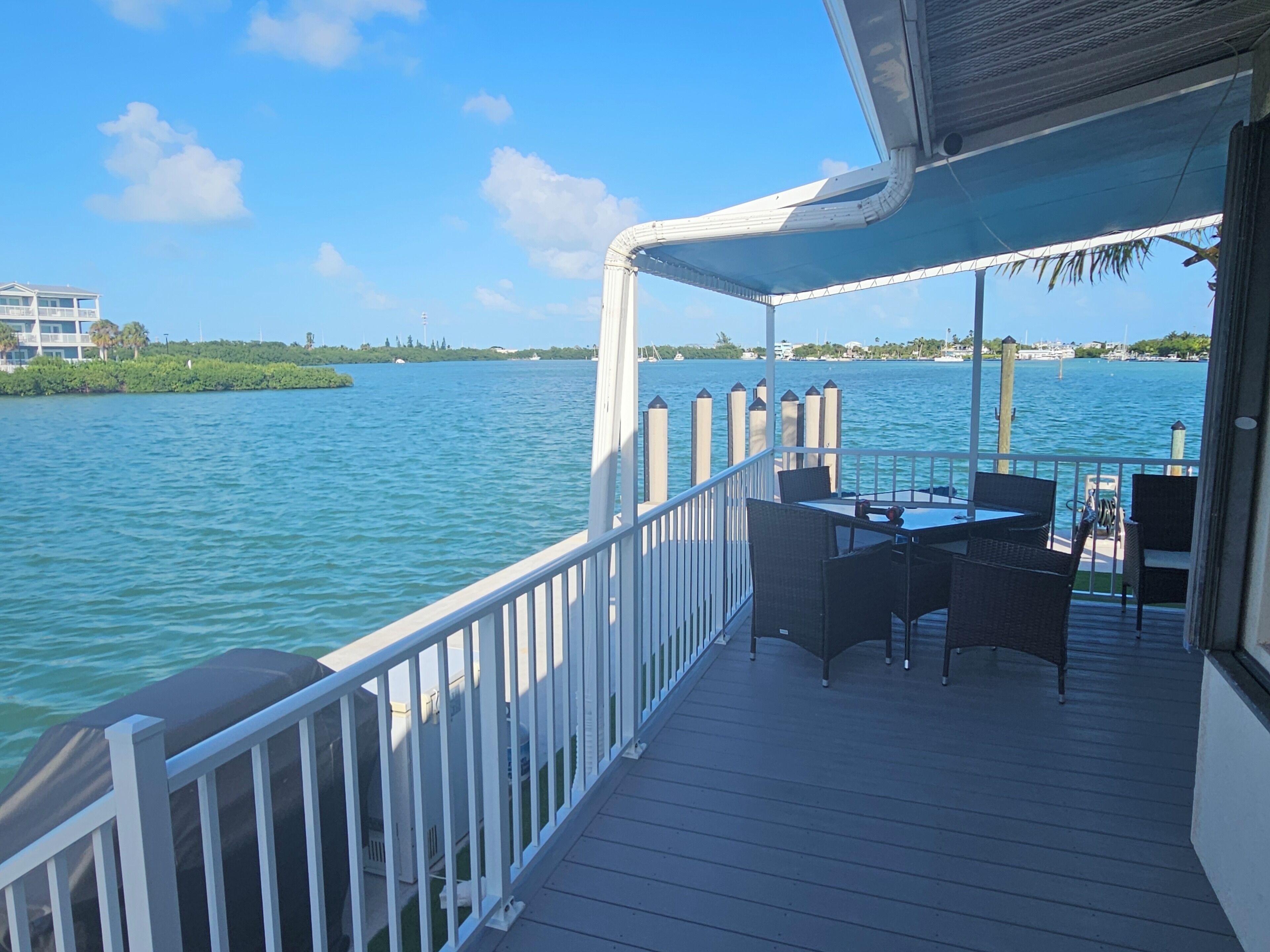 A deck with outdoor furniture overlooking a body of water, a railing, and a distant view of a building and trees.
