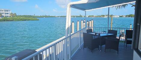 A deck with outdoor furniture overlooking a body of water, a railing, and a distant view of a building and trees.