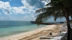 Ein Strand mit klarem türkisblauem Wasser, einem hölzernen Steg und Liegestühlen am Ufer.