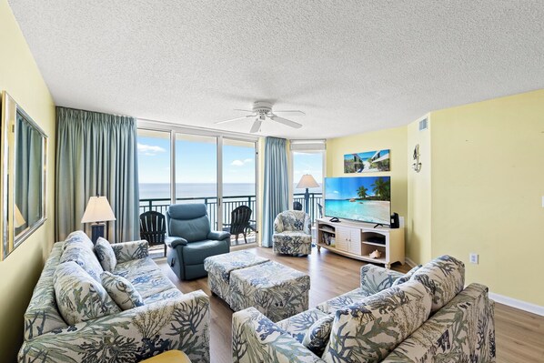 Een ruime woonkamer met een grote flatscreen-tv, comfortabele zithoek en uitzicht op de oceaan door grote ramen.