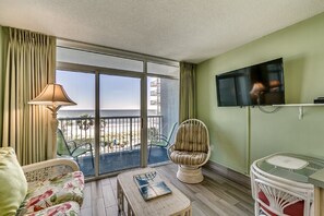 A balcony with a view of the beach, a television mounted on the wall, and a wicker chair.