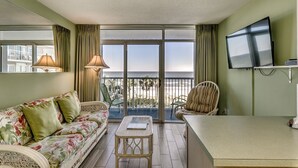 A balcony with a view of the beach, a sofa, a chair, a table, and a TV. - Tropical 3rd Floor Unit, Indoor and Outdoor Amenities | Sea Watch S - 311 (Myrtle Beach)