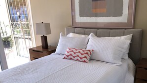 A neatly made bed with white bedding and a single patterned pillow, a bedside table with a lamp, and a framed abstract artwork above the bed. - Location, location, location!  El Paseo Jewel in the desert  (Palm Desert)