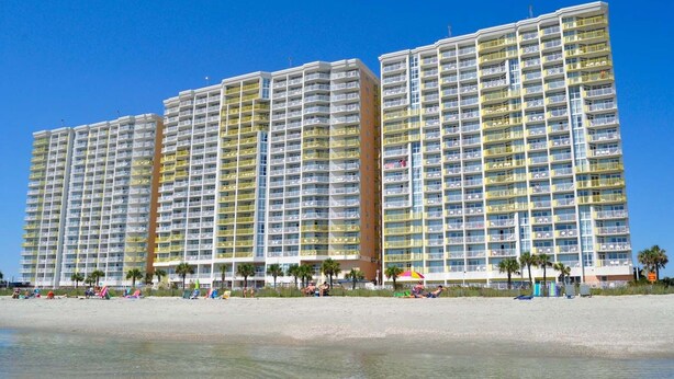 Un hôtel situé en bord de mer, composé de plusieurs immeubles de grande hauteur, doté de balcons et d’une plage de sable où l’on peut voir des gens et des chaises longues.
