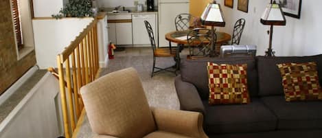 A living room with a sofa, armchair, and dining table. There is a kitchen in the background with white cabinets and a refrigerator.