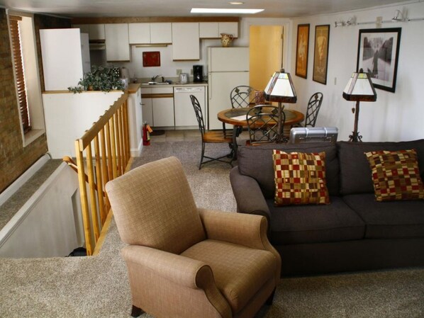 A living room with a sofa, armchair, and dining table. There is a kitchen in the background with white cabinets and a refrigerator.