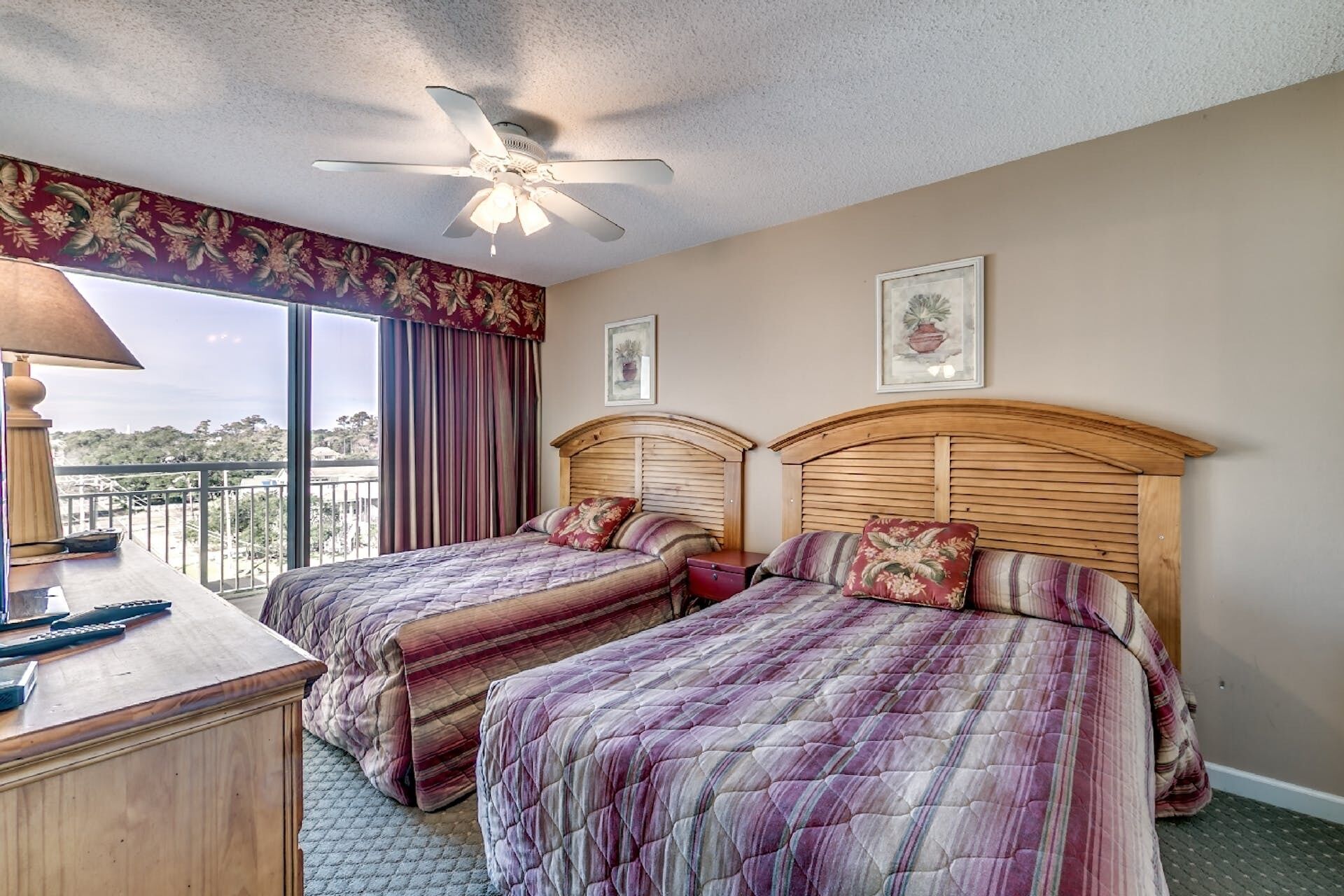 A hotel room with two beds, a ceiling fan, a desk with a lamp, and a view of the outdoors.