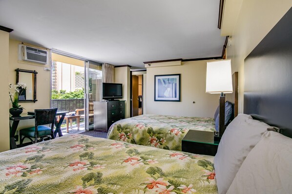 Two queen beds with floral quilts face a dark wood dresser and TV, next to sliding glass doors leading to a balcony.