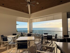 Een zonnig buitenterras met tafels en stoelen, met uitzicht op een strand en de oceaan.