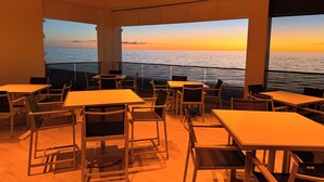 Un espace repas avec des tables et des chaises, de grandes fenêtres offrant une vue sur l’océan au coucher du soleil.