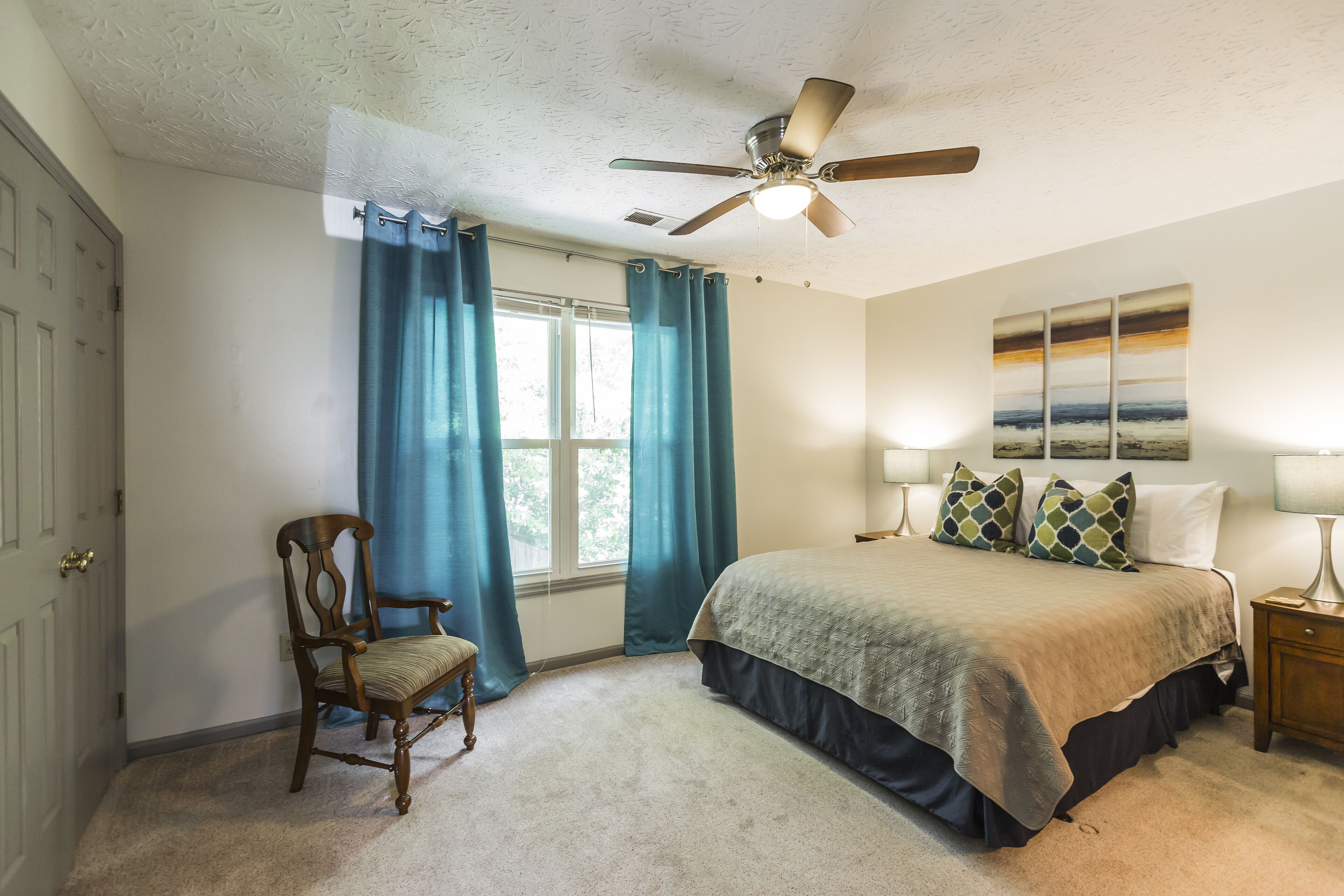 A bedroom with a bed, a chair, a ceiling fan, a window with curtains, and a painting on the wall.