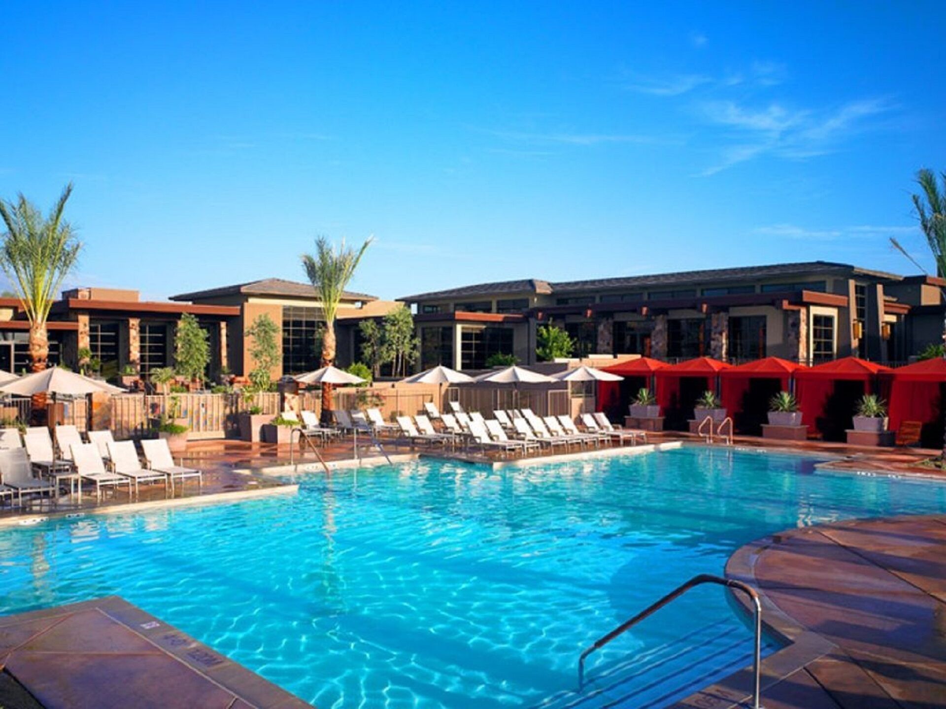 Ein Swimmingpool mit Liegestühlen und Sonnenschirmen, umgeben von Gebäuden mit großen Fenstern und Palmen.