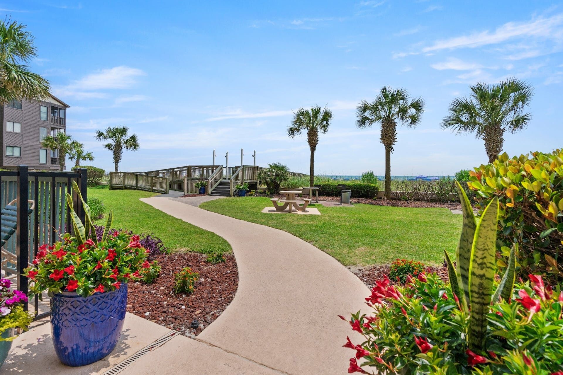 A pathway leading to a wooden bridge, surrounded by palm trees and flower beds.