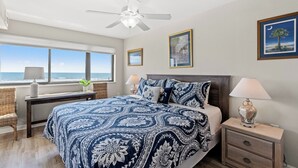 Ein Schlafzimmer mit Bett, Nachttischen, Deckenventilator, Schreibtisch und Fenster mit Meerblick.