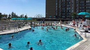 A pool area with multiple people swimming and lounging, surrounded by a building with balconies. - Bethany Beach • Lakefront Sea Colony Condo • Walk to Beach • Sleeps 8 (Bethany Beach)