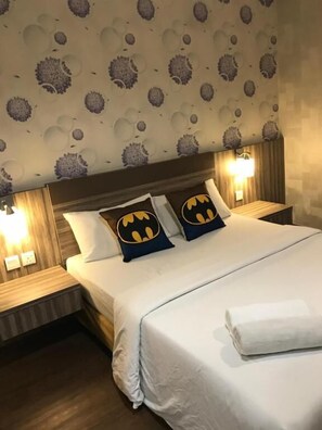 A neatly made bed with white bedding and two pillows featuring a Batman logo. The room has a patterned wallpaper and two bedside lamps. - Victory Hotel (Johor Bahru)