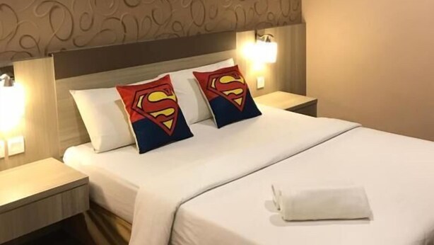 A hotel room with a patterned wallpaper, a bed with white bedding, two Superman-themed pillows, and bedside lamps. - Victory Hotel (Johor Bahru)
