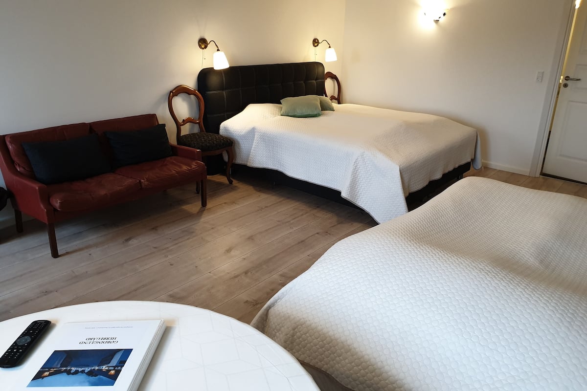 A hotel room with a bed, a sofa, a chair, and a bedside table with a book.