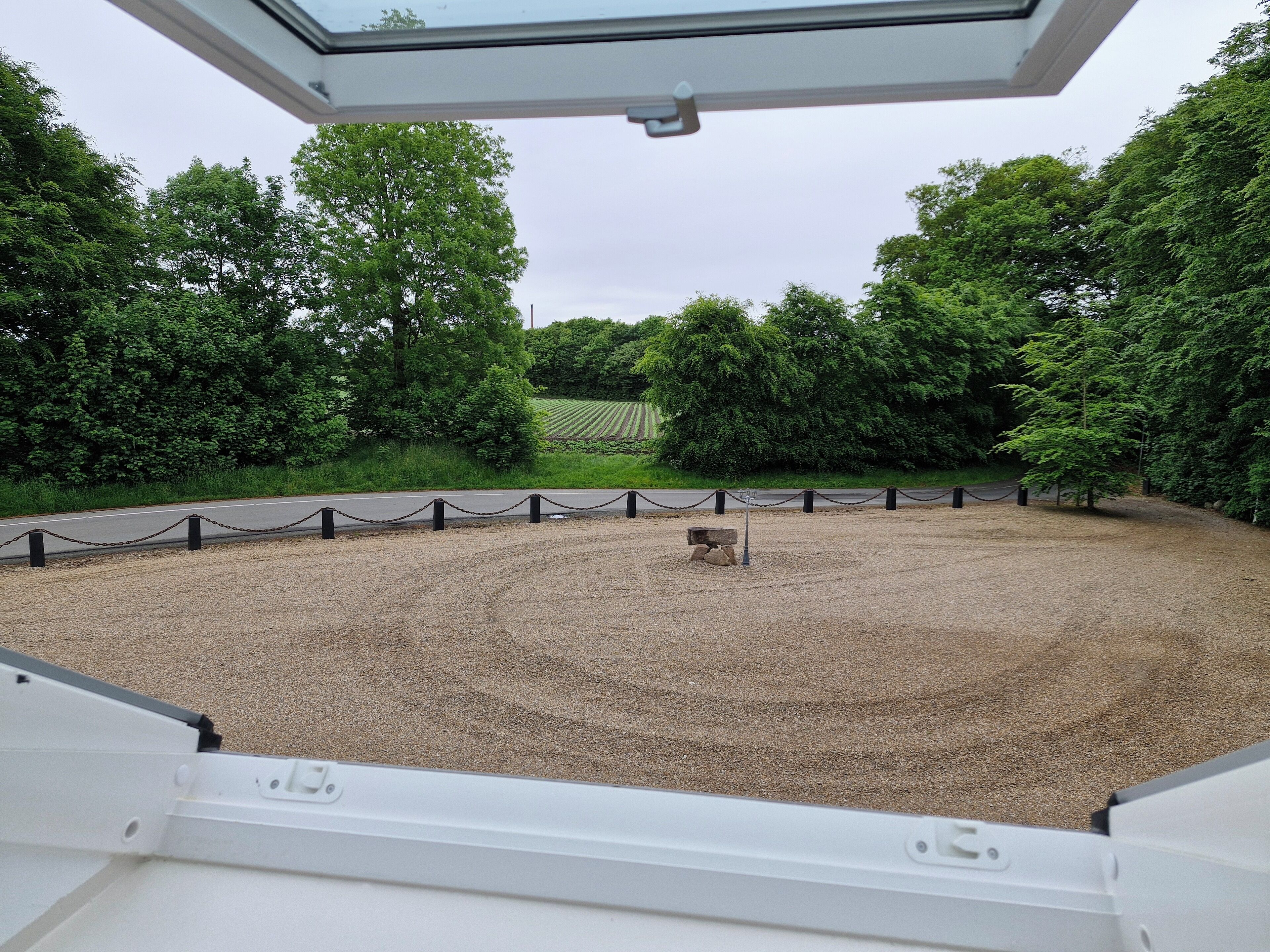 A view from a window overlooking a gravel area with a stone bench, surrounded by trees and a chain-link fence.