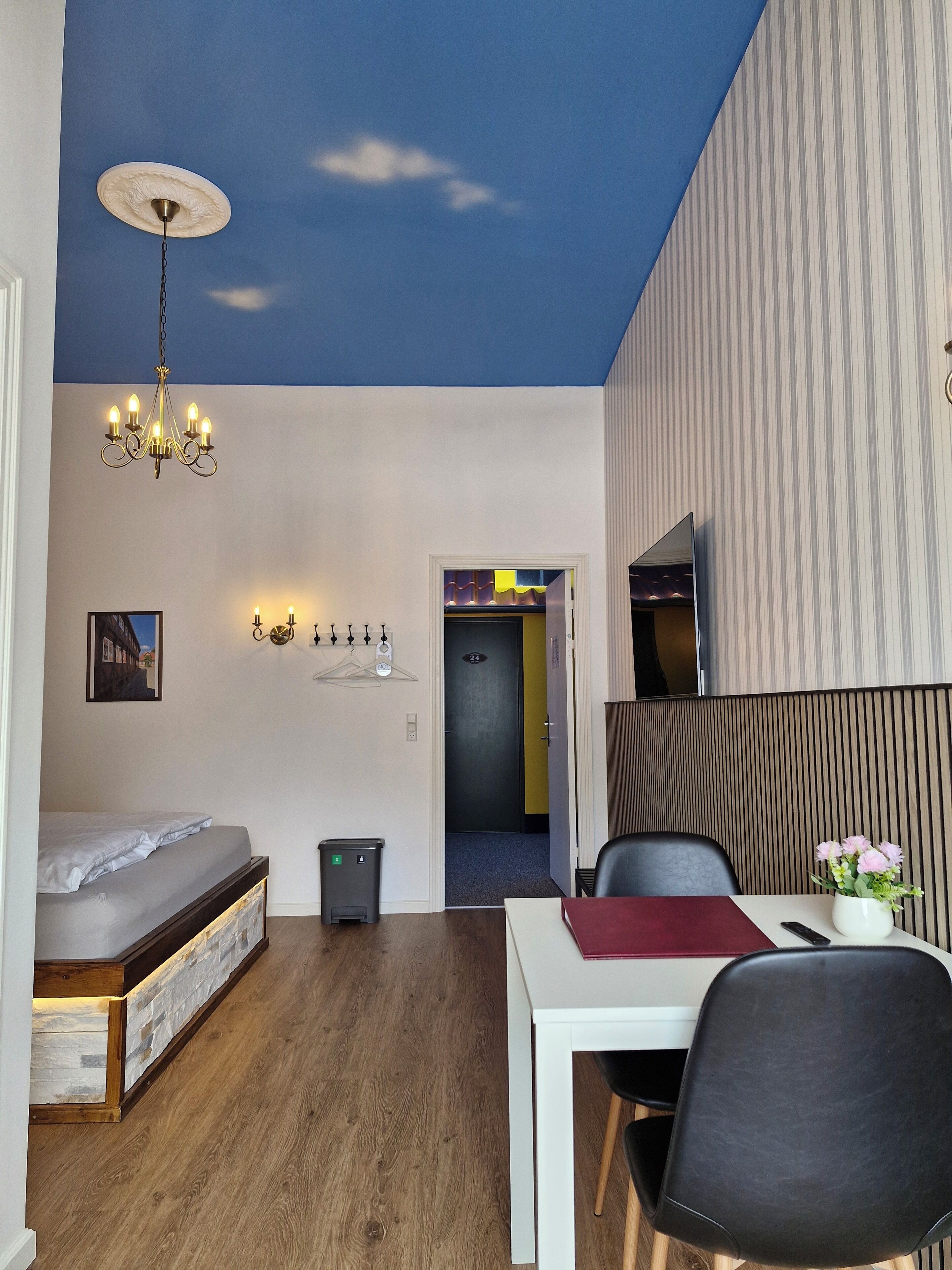 A compact hotel room with a bed, a desk with a chair, a chandelier, a wall-mounted TV, and a door leading to another room.
