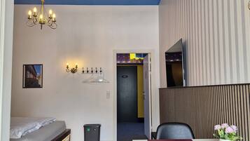 Una camera d'albergo compatta con un letto, una scrivania con una sedia, un lampadario, un televisore montato a parete e una porta che conduce a un'altra stanza.