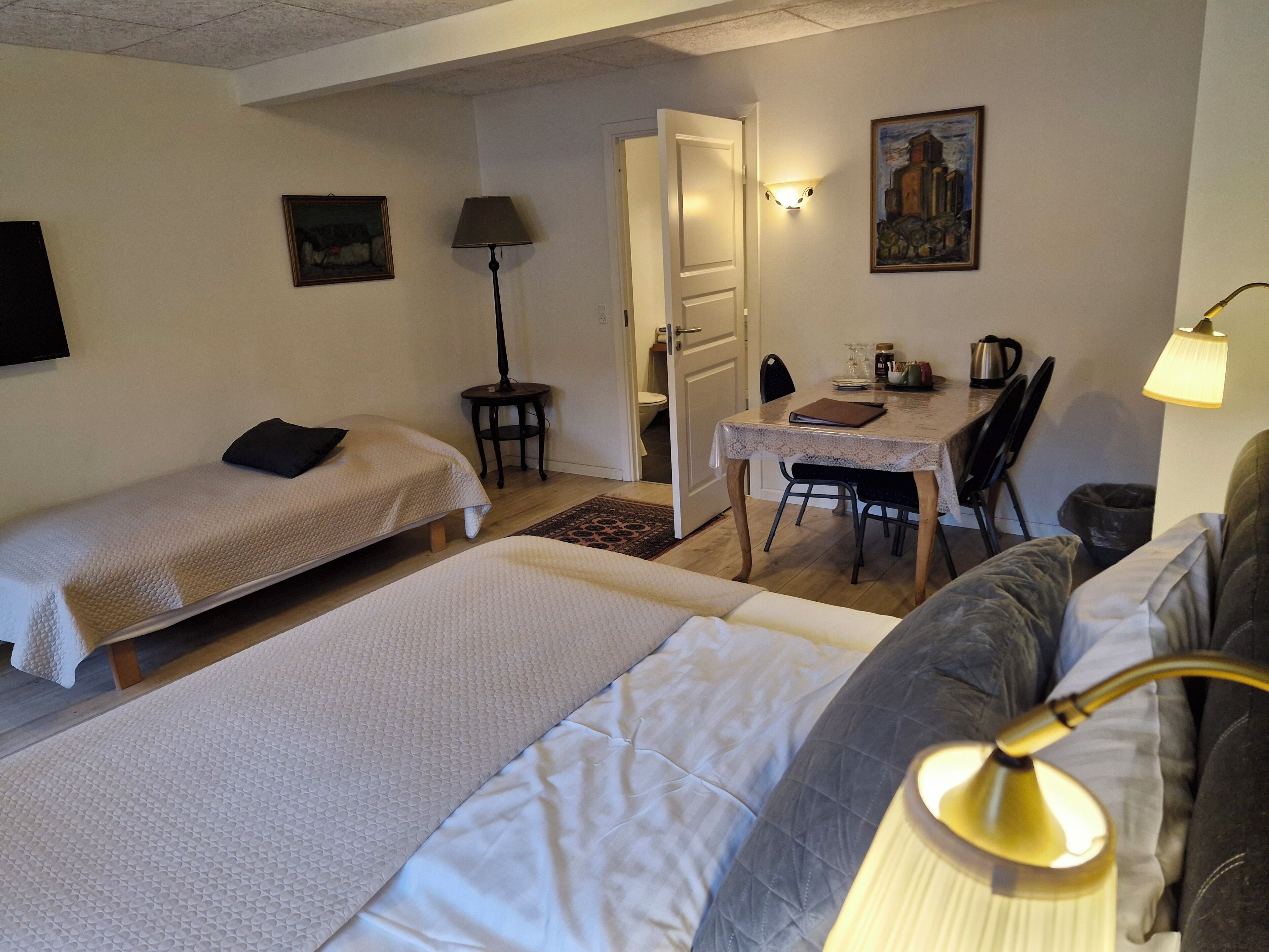 A room with two beds, a dining table with chairs, a lamp, and a painting on the wall.