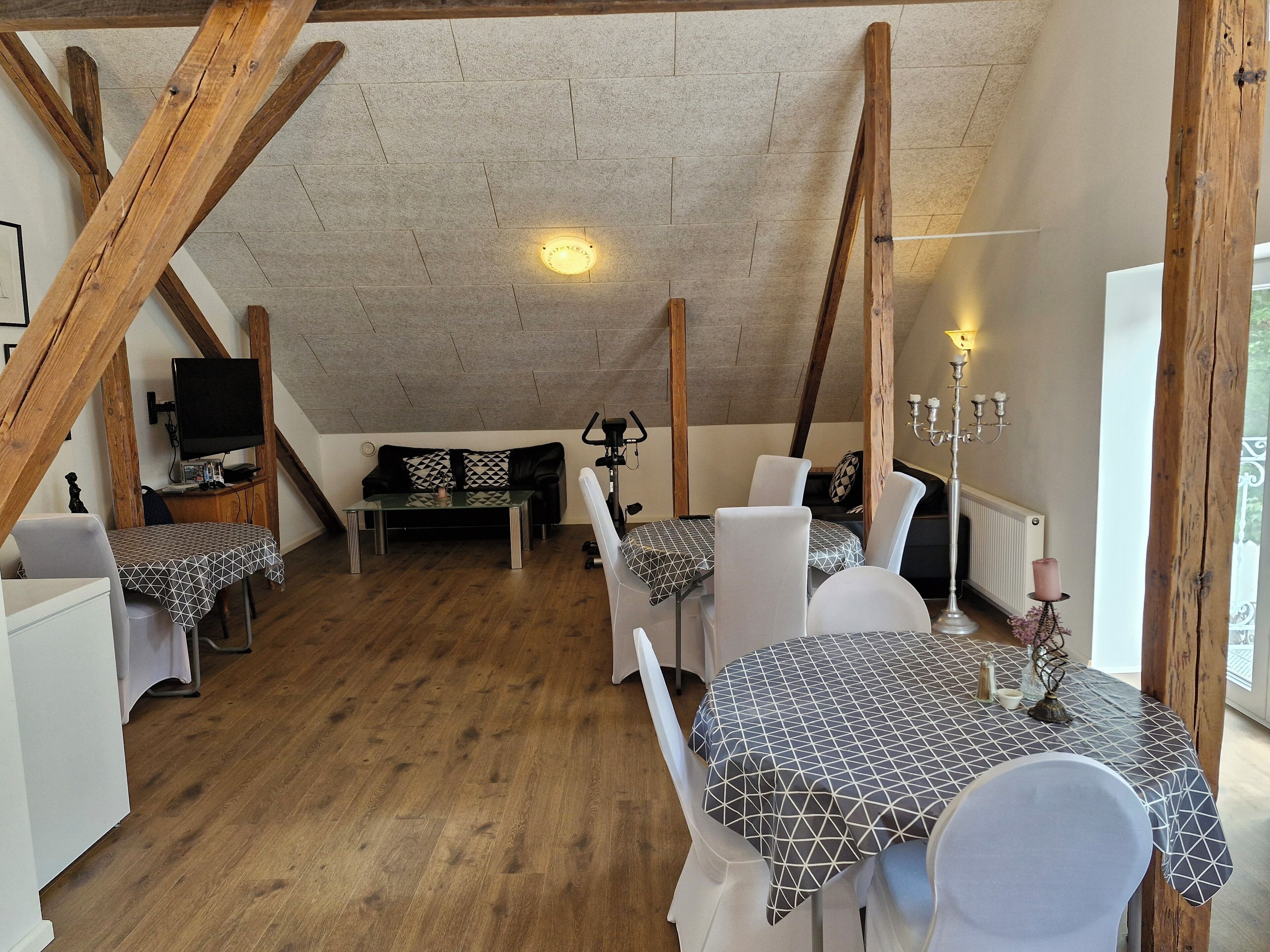 Ein gemütliches Dachzimmer mit freiliegenden Holzbalken, einem Schlafplatz in der Schräge, einem Esstisch mit Stühlen, einem Fernseher und einem Kerzenständer.
