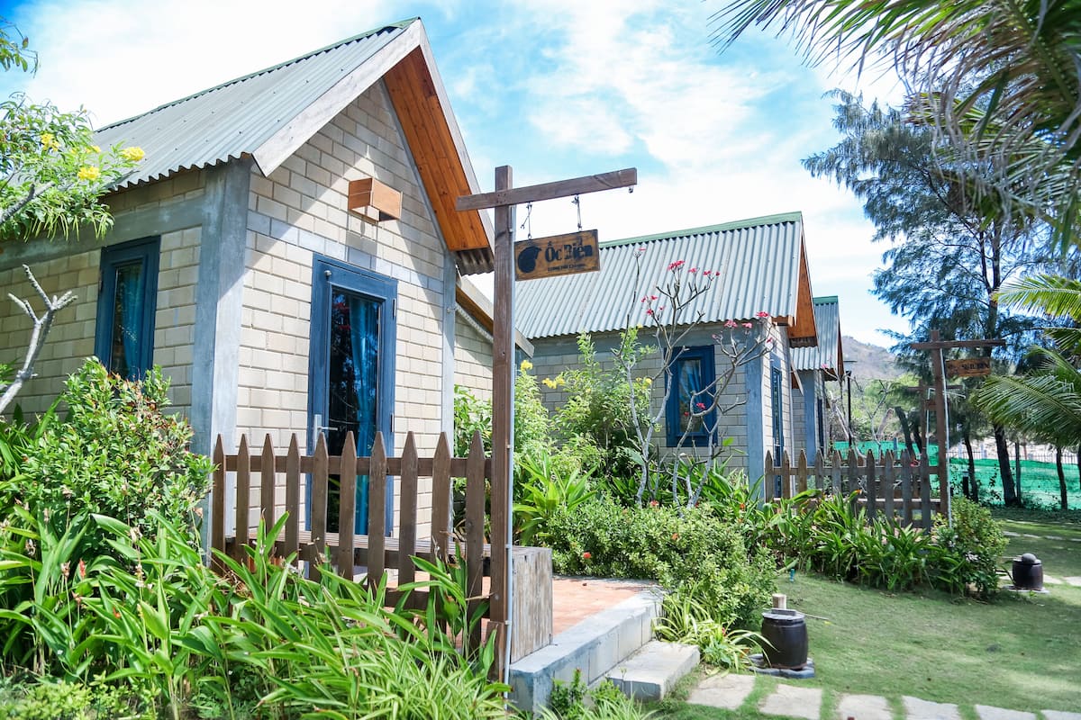A rustic wooden house with a corrugated metal roof, surrounded by lush greenery and a wooden fence.