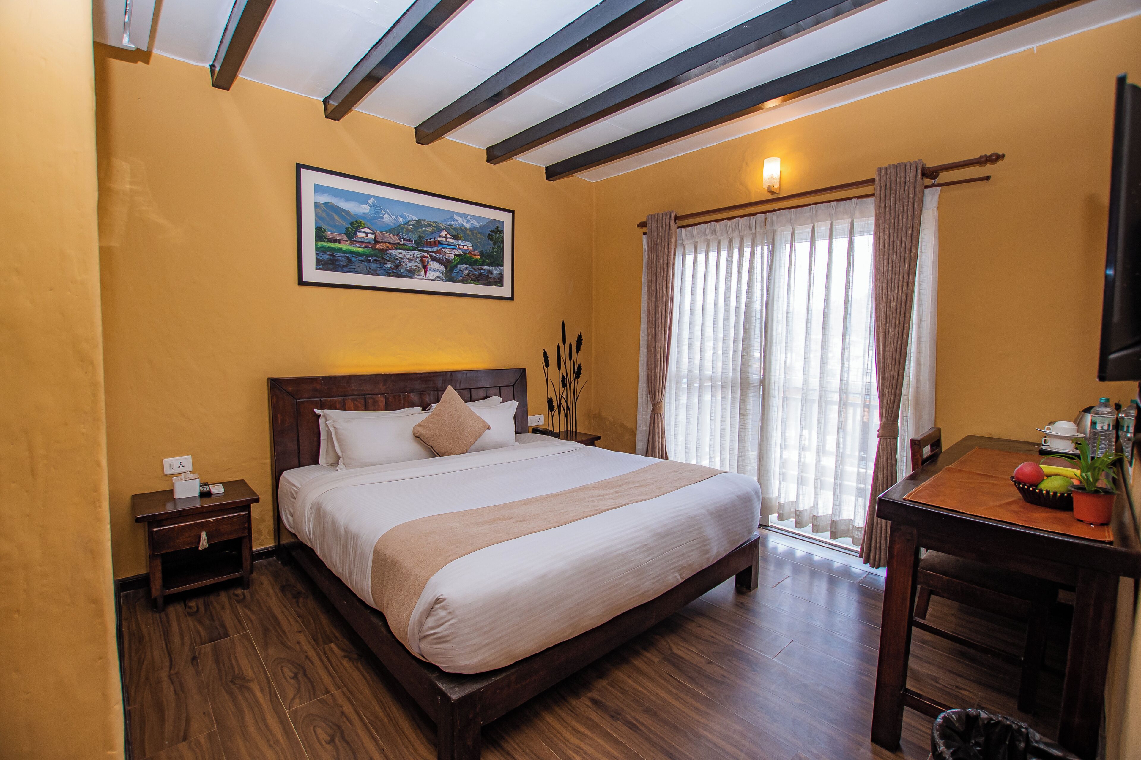 A hotel room with a wooden bed, a nightstand, a desk with a plant, and a painting on the wall.