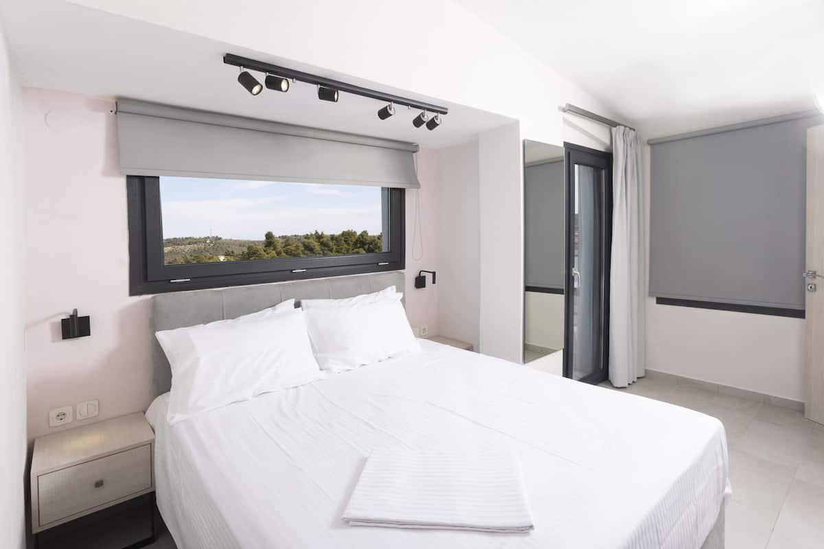 A modern hotel room with a large bed, a window with a view, a bedside table, and a door leading to a balcony.