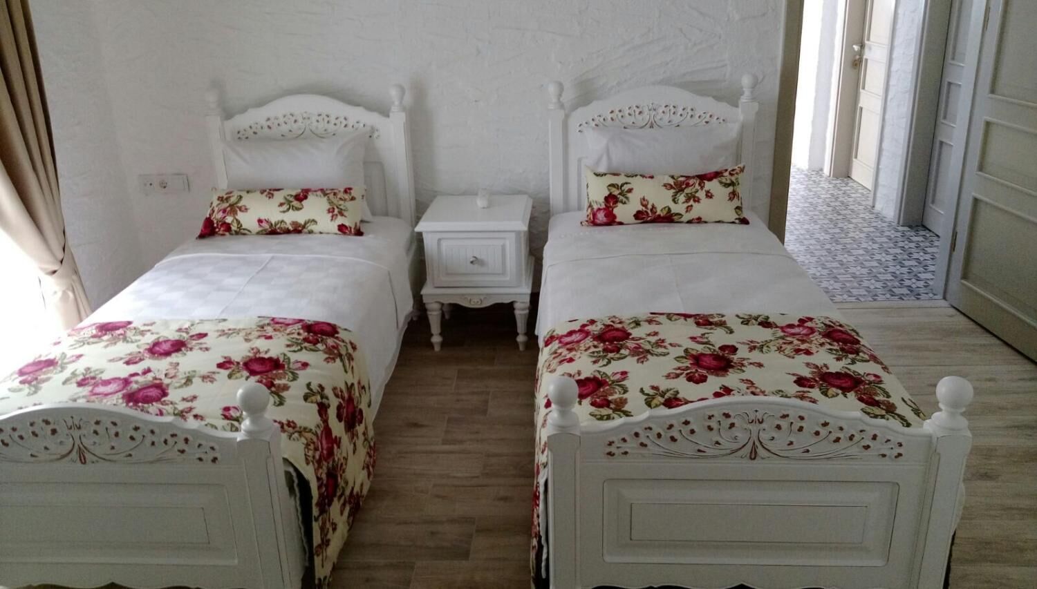 A double room with two single beds, each with a floral bedspread and matching pillows. There are bedside tables with drawers, and the room has a tiled floor.