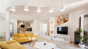 Un salon moderne avec un canapé, une table basse et un téléviseur. On aperçoit une cuisine au fond et un escalier menant à un autre niveau.