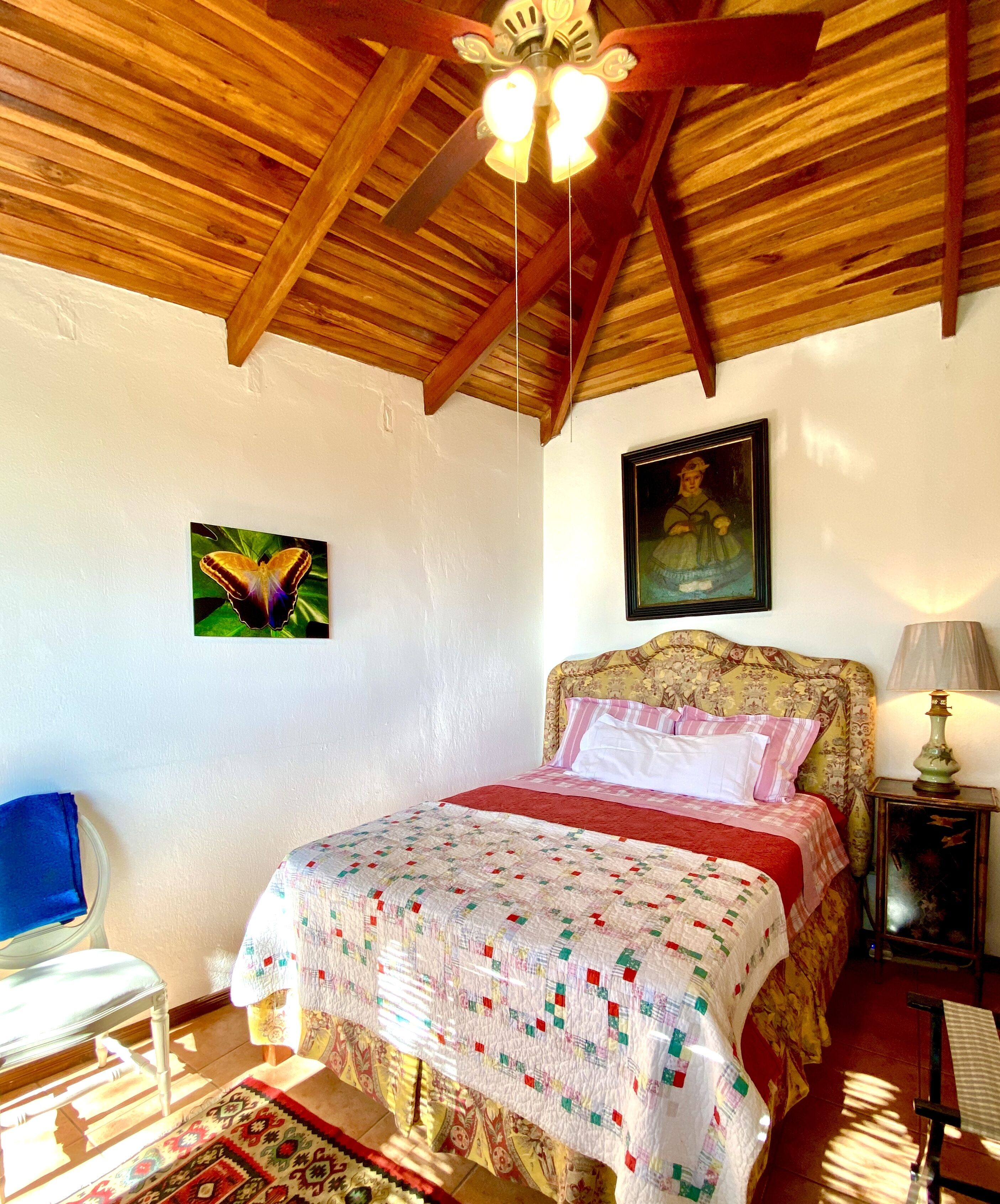 Une chambre avec un plafond en bois, un lit doté d’une tête de lit à motifs floraux, un tableau accroché au mur et une chaise avec un coussin bleu.