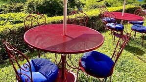 Une terrasse avec des tables en métal rouge et des chaises bleues, des parasols blancs, et une vue sur des collines et des montagnes verdoyantes.