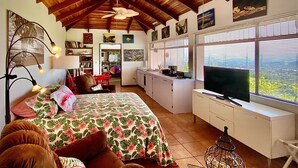 Une pièce spacieuse avec un plafond haut, un lit recouvert d’une literie à motifs floraux, un téléviseur posé sur un meuble et une vue dégagée grâce à de grandes fenêtres.