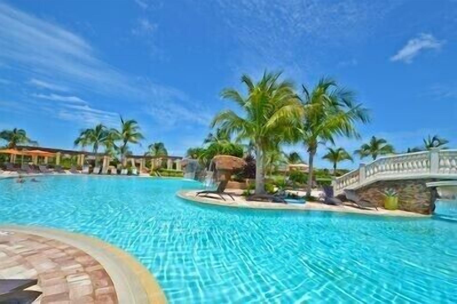 A large outdoor swimming pool with clear blue water, surrounded by palm trees and a stone bridge.